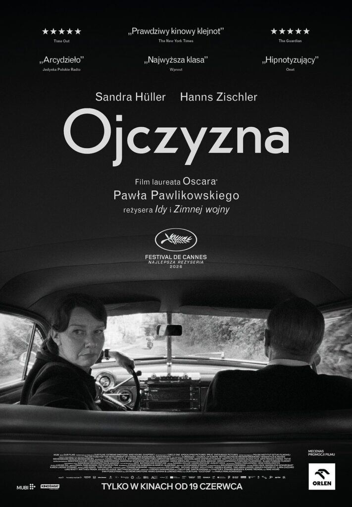 Dwoje dorosłych osób: kobieta i mężczyzna siedzą w samochodzie. Kobieta prowadzi auto, trzyma lewą ręką kierownicę, odwraca się do tyłu. Droga prowadzi wśród drzew. 