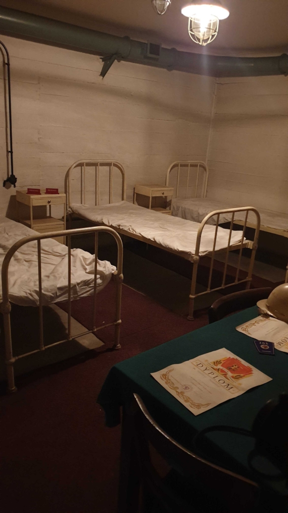 Zdjęcie schronu. Trzy metalowe łóżka ustawione obok siebie, w prawym dolnym rogu widoczny blat stołu na którym leżą dyplomy