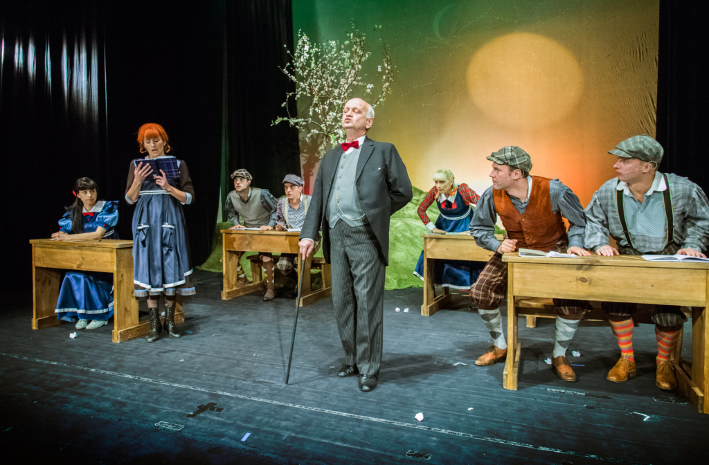 Na scenie teatralnej ustawione są drewniane ławki szkolne, przy których siedzą aktorzy w kolorowych, stylizowanych na dawne czasy strojach. Po lewej dziewczynka w granatowej sukience z czerwoną kokardą patrzy w bok, obok niej rudowłosa aktorka w niebieskiej sukni czyta z tabliczki. W centrum starszy mężczyzna w eleganckim garniturze z czerwonym muszkiem i laską stoi w autorytatywnej pozie. Po prawej stronie kilku uczniów w czapkach i kamizelkach pochyla się nad swoimi ławkami, jeden z nich nosi w paski skarpety w jaskrawych kolorach. W tle widoczny jest kwitnący gałązkami rekwizyt drzewa oraz malowana sceneria przypominająca krajobraz z zachodzącym słońcem.