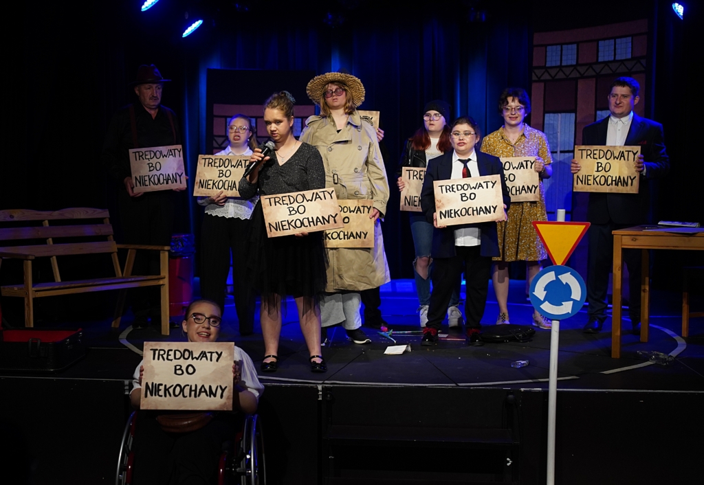 Grupa aktorów na scenie podczas spektaklu, trzymających tabliczki z napisem „Tredowaty, bo niekochany”. W tle scenografia teatralna - stół, ławka, obrysy bloków mieszkalnych. Przed sceną po lewej stronie jeden z aktorów na wózku inwalidzkim, a po prawej znaki drogowe wjazdu na rondo.