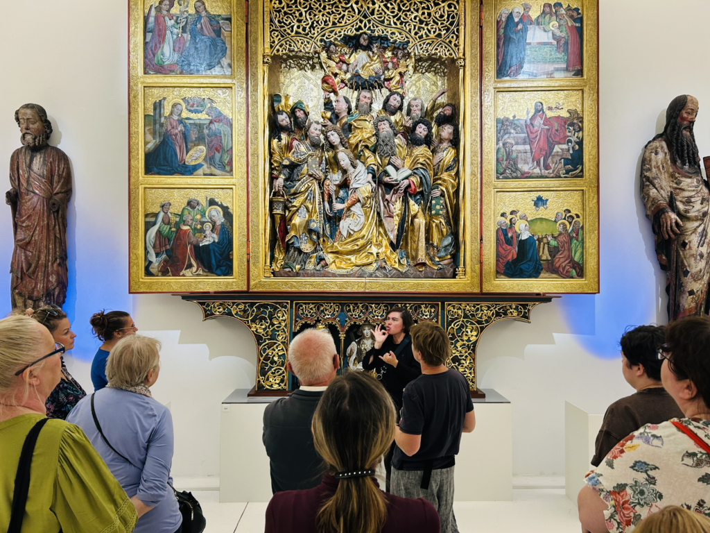 Fotografia wykonana we wnętrzu muzealnym o jasnych ścianach. Na pierwszym planie widać grupę zwiedzających – kobiety i mężczyzn w różnym wieku, stojących tyłem i półprofilem do obiektywu. W centrum kadru stoi przodem do grupy tłumaczka PJM, unosząca dłoń w wyrazistym geście przy twarzy.  Tło kadru tworzy wielki, otwarty ołtarz szafiasty w bogato zdobionej, złoconej oprawie. W jego głębokiej kwaterze środkowej znajduje się rzeźbiona, wielopostaciowa scena Zaśnięcia Marii, a na otwartych skrzydłach bocznych widoczne są prostokątne kwatery z malowanymi scenami - po 3 na każdym skrzydle. Całość spoczywa na dekoracyjnej predelli. Po lewej i prawej stronie ołtarza, stoją wysokie, polichromowane figury apostołów rzeźbione w drewnie.