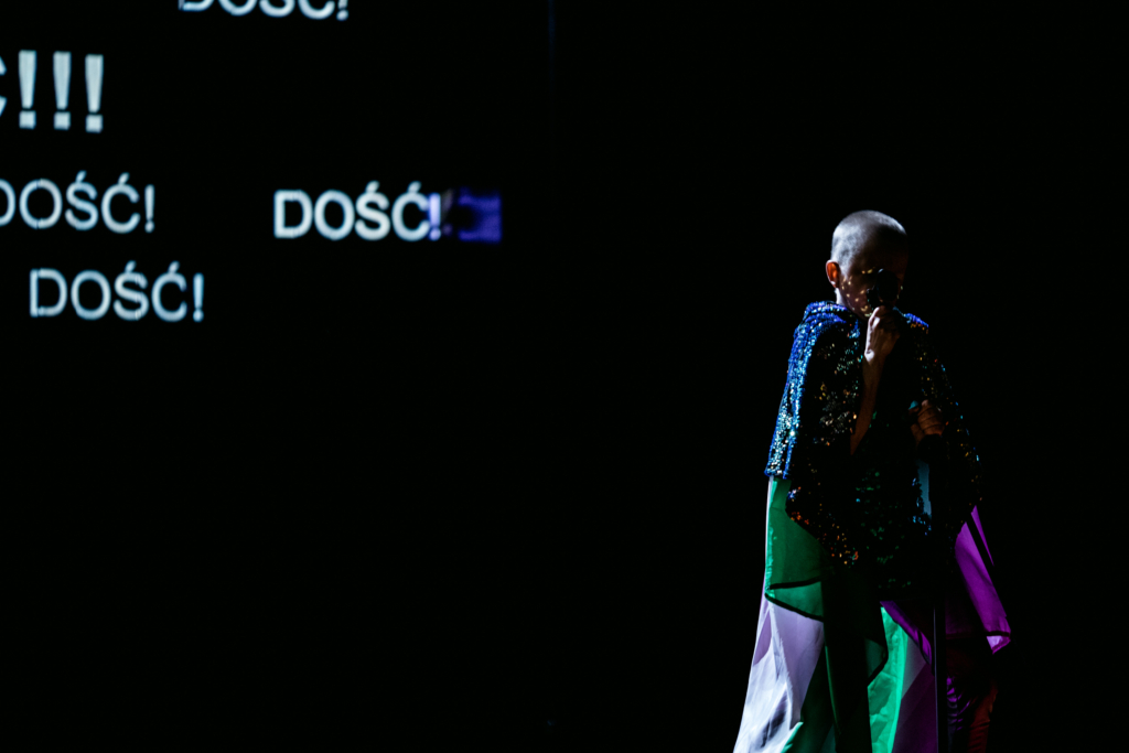 Na ciemnej scenie stoi performer w błyszczącej, wielobarwnej pelerynie i trzyma mikrofon przy twarzy. Na czarnym tle wyświetlają się duże białe napisy „DOŚĆ!”. Oświetlenie sceniczne podkreśla sylwetkę performera intensywnymi zielonymi, fioletowymi i niebieskimi refleksami.