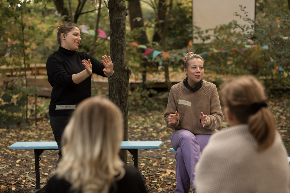 Zdjęcie: grupa osób podczas warsztatów improwizacji teatralnej w plenerze. Trzy osoby siedzą na ławkach, jedna osoba tłumaczy na polski język migowy.