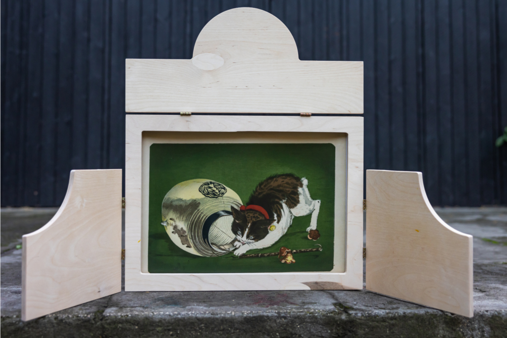 Zdjęcie: Drewniana rama do teatrzyku kamishibai z ilustracją przedstawiającą kota i lampion. Tło: ściana drewnianego domku na Osiedlu Jazdów.