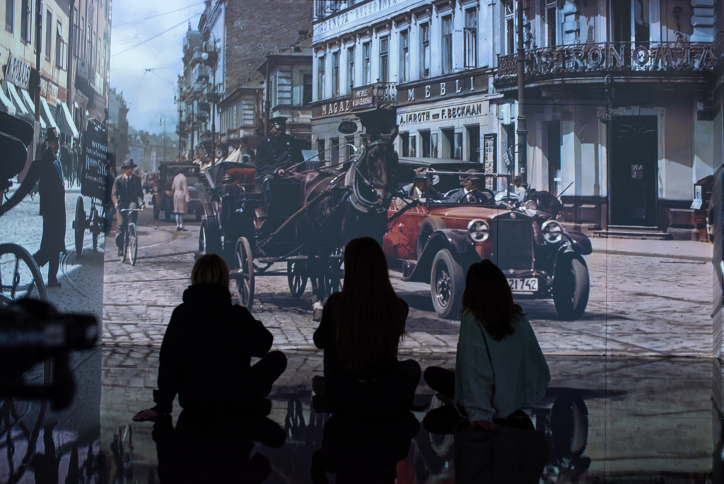 Zdjęcie z wystawy. Wielkoformatowa fotografia ulicy starej Warszawy. Przedstawia konny powóz i stare auto na tle budynku. Przed fotografią na ławce, siedzą trzy osoby.