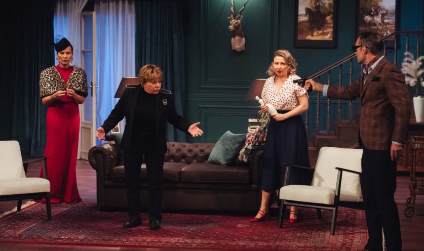 Na fotografii widoczna jest scena teatralna w eleganckim, stylizowanym salonie z ciemnozielonymi ścianami, obrazami, schodami, sofą i ozdobnym dywanem. Po lewej stronie stoi kobieta w długiej czerwonej sukni i leopardziej narzutce. Obok niej osoba w czarnym stroju rozkłada ręce w teatralnym geście. Po prawej stronie znajduje się blondynka ubrana w białą bluzkę w czerwone kropki oraz granatową spódnicę. Trzyma w dłoniach jasny przedmiot i patrzy z niepokojem przed siebie. Skrajnie po prawej stoi mężczyzna w brązowej kraciastej marynarce i okularach, który wyciąga rękę z pistoletem skierowanym w stronę pozostałych osób. Scena ma charakter przedstawia napiętą, kryminalną sytuację pomiędzy osbami. 